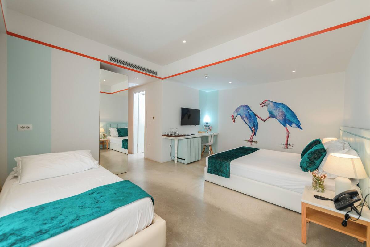 A hotel room with two beds, a desk, a TV, and a mural of birds.