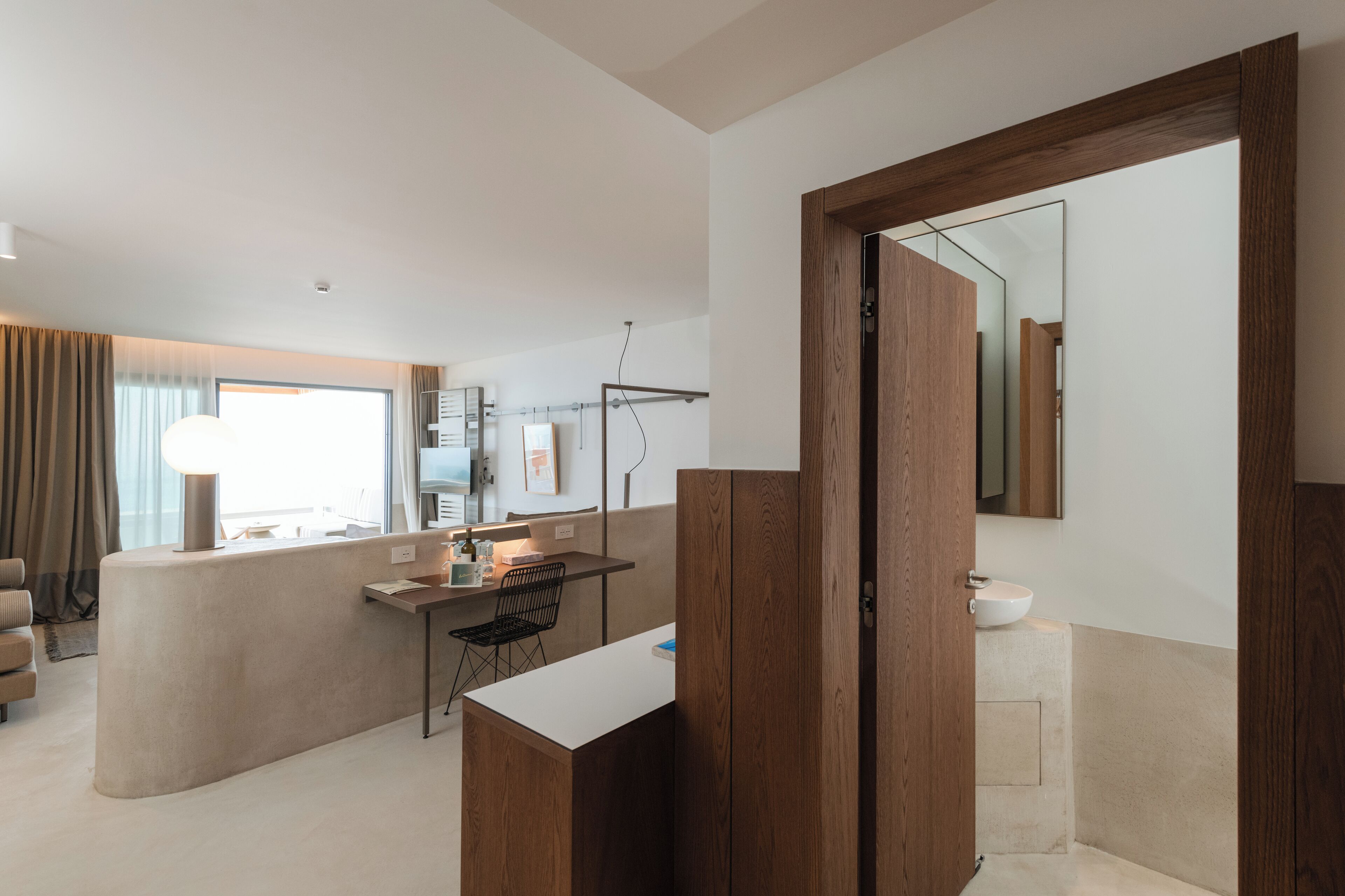 A modern hotel room with a large window, a wooden vanity, a desk, and a chair.
