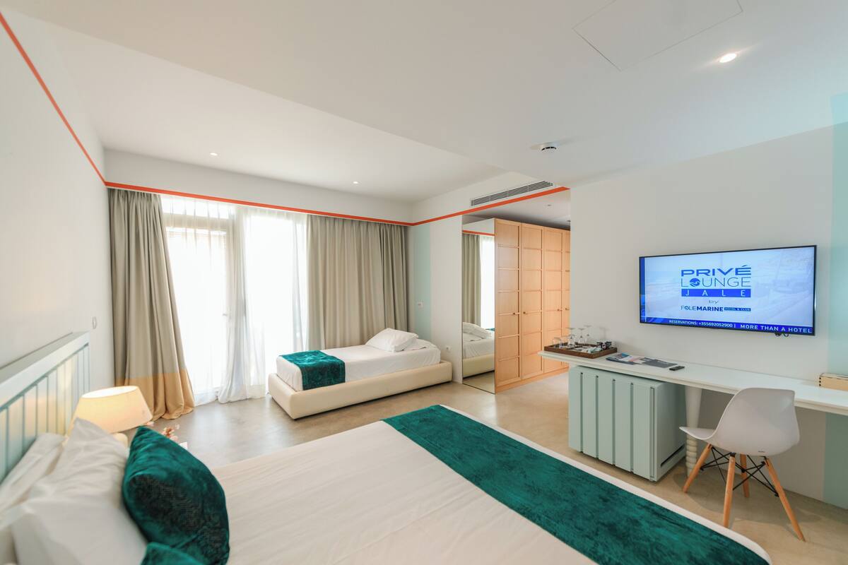 A modern hotel room with a bed, a desk, a TV, and a view of the sea.