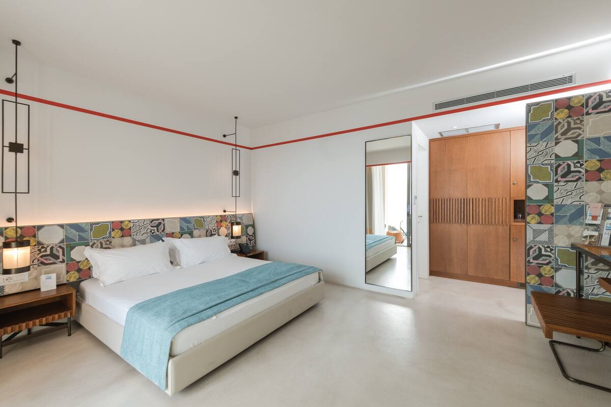 A modern hotel room with a large bed, bedside tables, a mirror, and a wooden wardrobe.