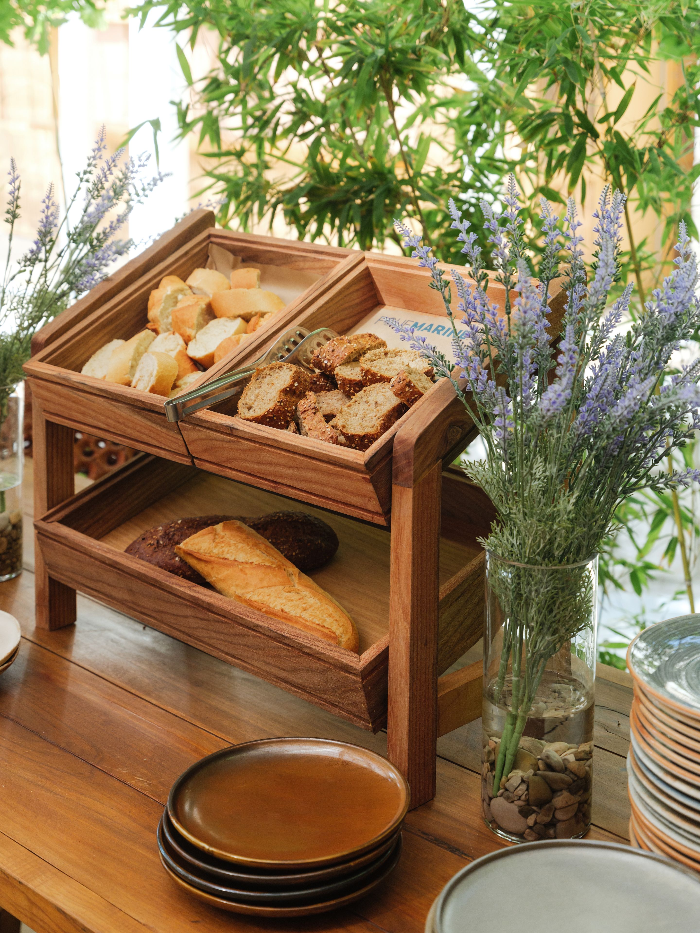 A wooden bread display with various types of bread, a vase with lavender, and stacked plates on a wooden table.