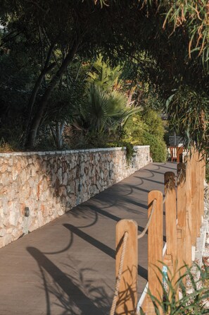 A paved pathway bordered by a stone wall and wooden fence, leading through a lush garden with various plants and trees. - Folie Marine Beach Hotel & Club (Himare)