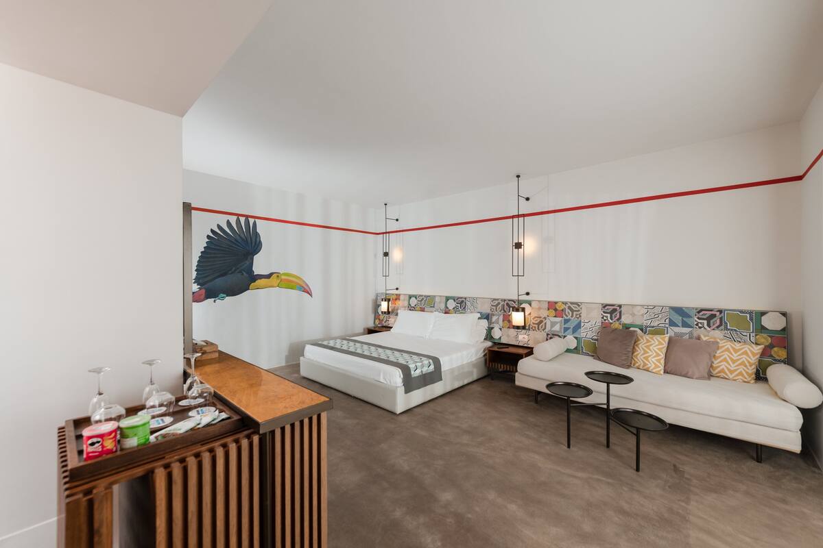 A modern hotel room with a large bed, a sofa, a small table, and a wall-mounted artwork of a bird.