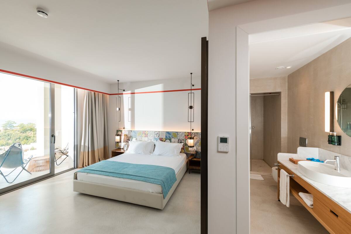 A modern hotel room with a large bed, a bathroom with a vanity, and a balcony with a chair.