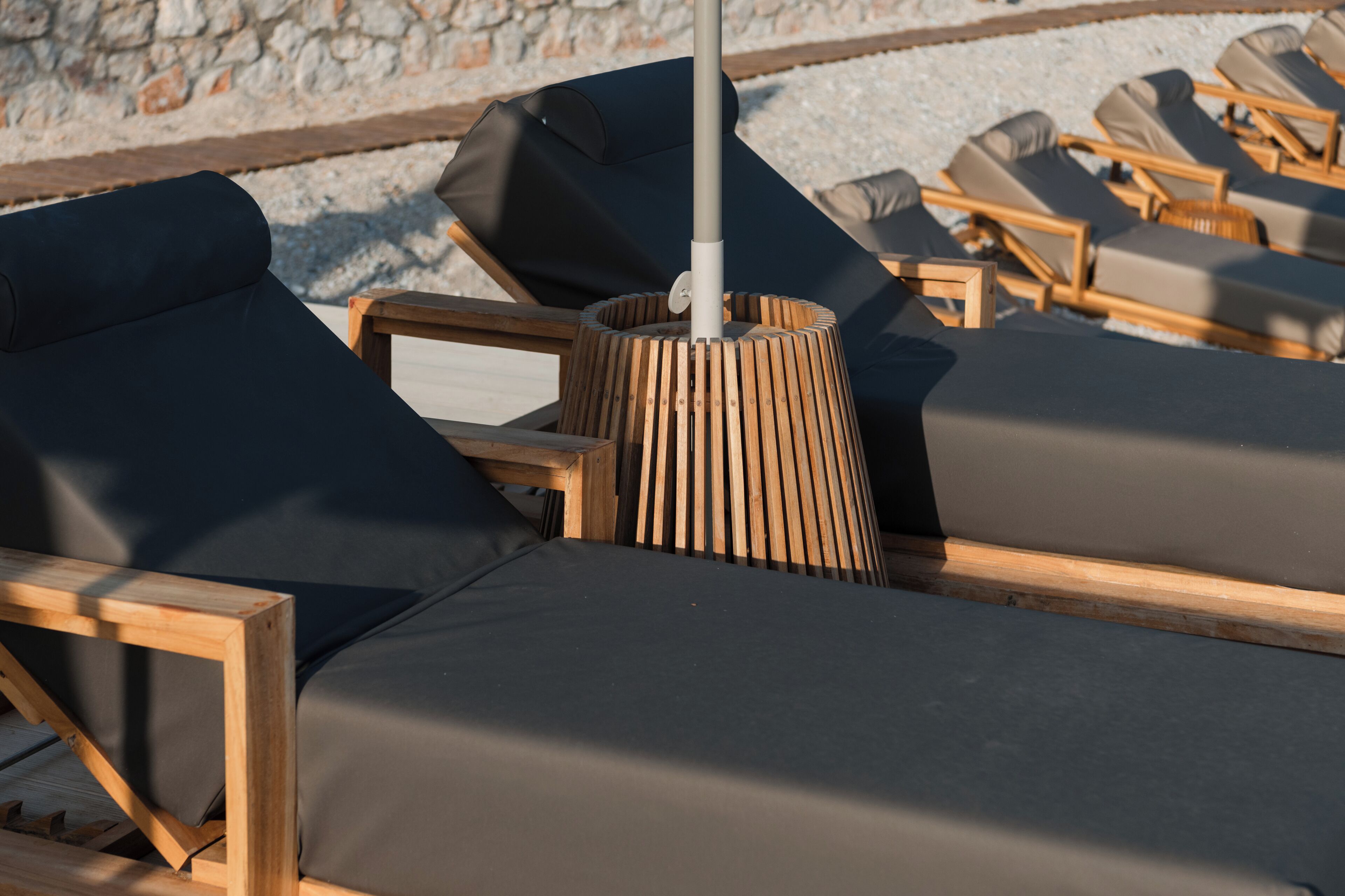 A row of wooden lounge chairs with dark cushions, arranged outdoors with a stone wall in the background.