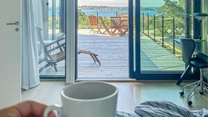 Een kamer met een grote glazen schuindeur die leidt naar een terras met uitzicht op de zee en een plaatsje in de verte.