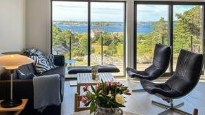 Een moderne woonkamer met een bank, een fauteuil, een tafel en een lamp. Door grote ramen is er uitzicht op zee en groen.