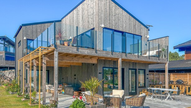 Een modern houten huis met twee verdiepingen, grote glasramen, buitenmeubels en een terras.