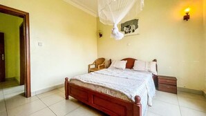 A bedroom with a wooden bed, a bedside table, a nightstand, a wall-mounted lamp, and a hanging mosquito net. - ibizza resort (Shingiro)