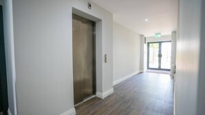 A hallway with a metallic elevator, white walls, and wooden flooring.