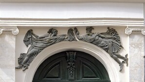 Een klassiek architectonisch detail met een reliëfbeeld van een rustende figuur boven een donkere, gewelfde deuropening.