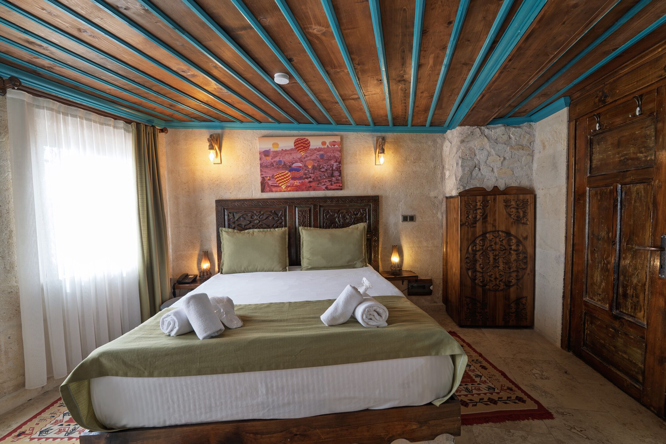 A hotel room with a large bed, wooden ceiling beams, a stone wall, and a wooden wardrobe.
