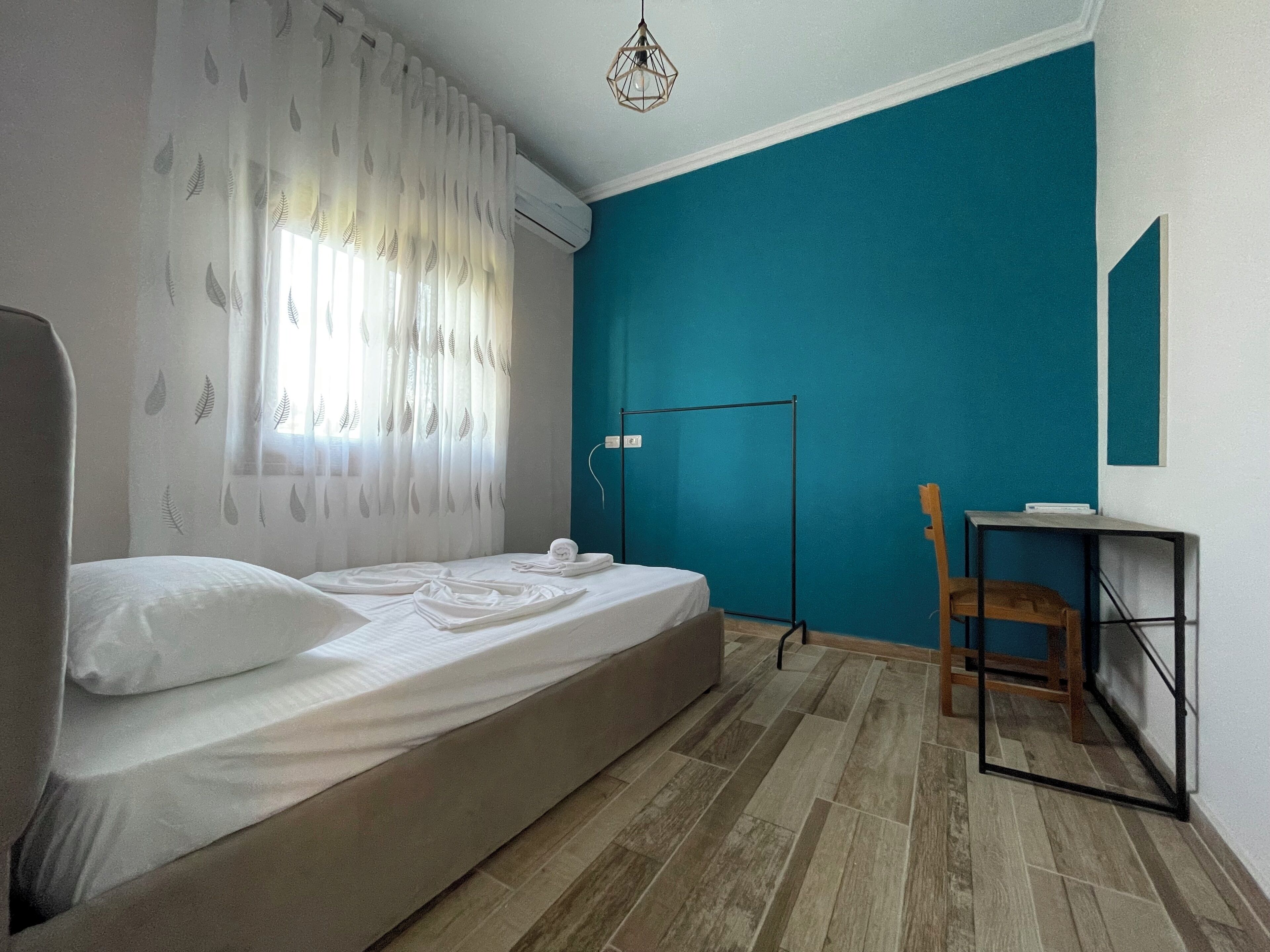 A bedroom with a bed, a desk, a chair, a standing lamp, and a window with sheer curtains.
