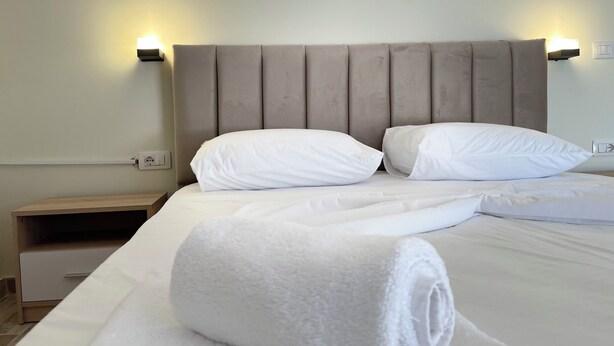 A neatly made bed with white linens and pillows, a wooden nightstand, and wall-mounted light fixtures.