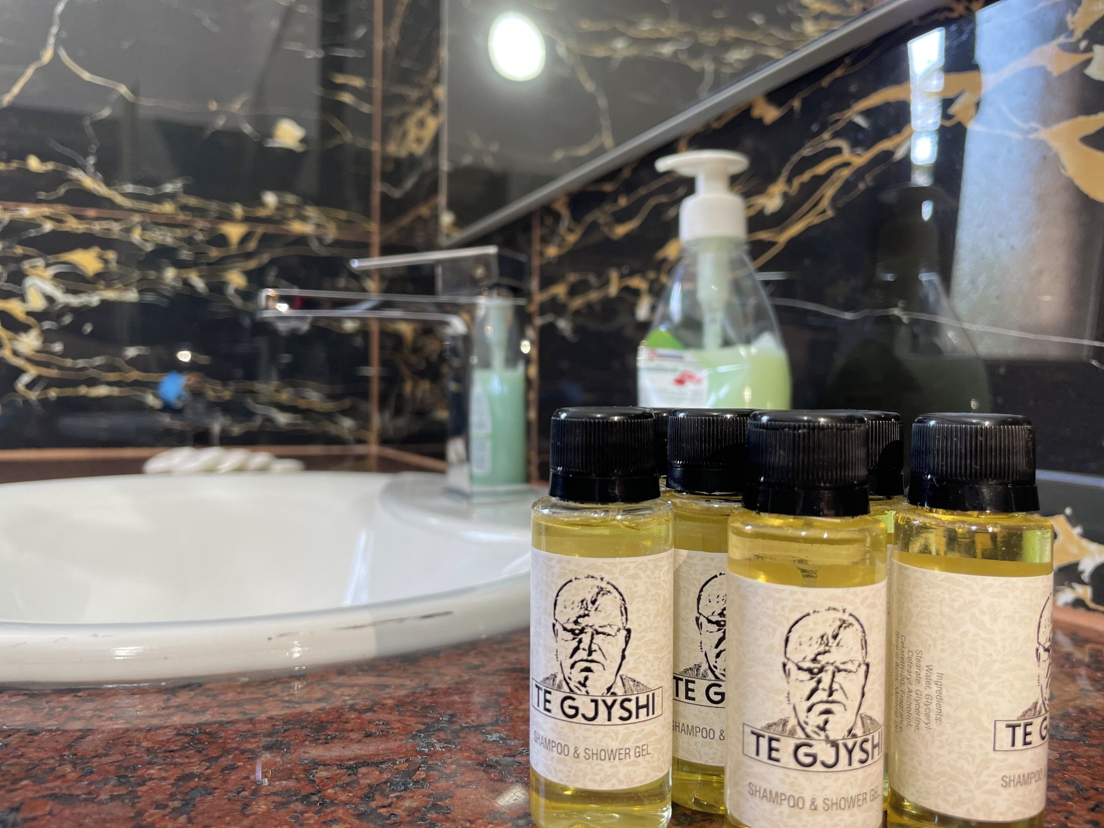 A bathroom with a marble sink, a mirror, and three bottles of shampoo and shower gel labeled "TE GJYSHI" on a granite countertop.