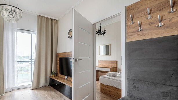A modern bedroom with a large bed, a TV, a wooden headboard, a bedside table, and a window with curtains. - RentPlanet - Bliska Wola (Warsaw)
