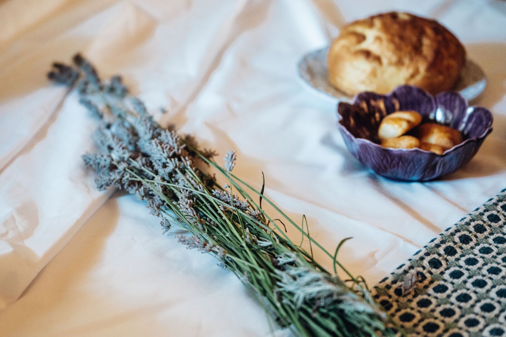 A bed with a white sheet, a bowl of cookies, and a bunch of dried lavender.