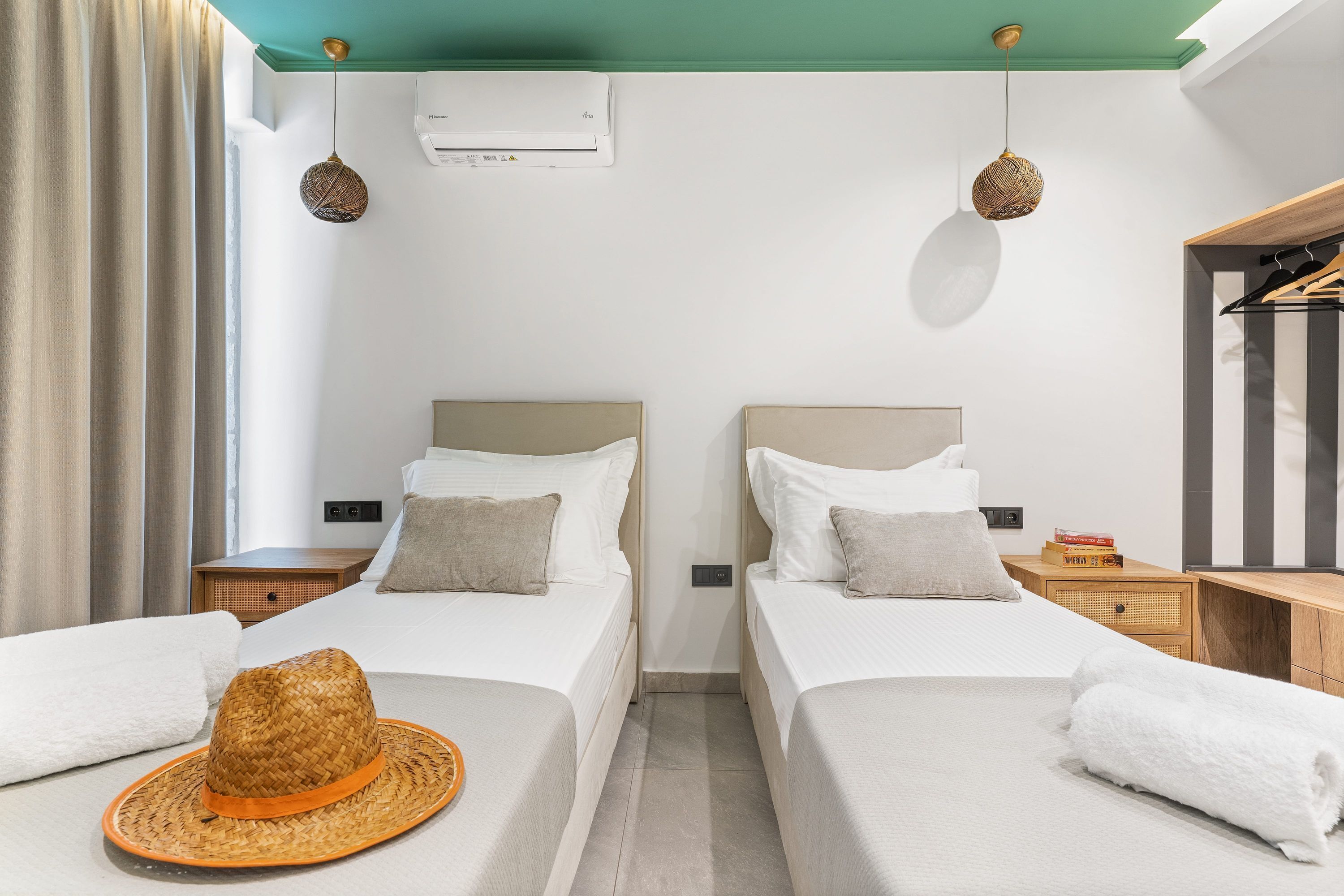 A hotel room with two beds, a ceiling fan, an air conditioning unit, a hat on one bed, and a wooden nightstand with a book.