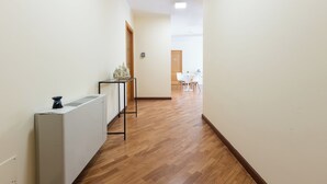 Un pasillo con un radiador, una pequeña mesa con un florero y una puerta que da a una habitación con una mesa y sillas blancas.