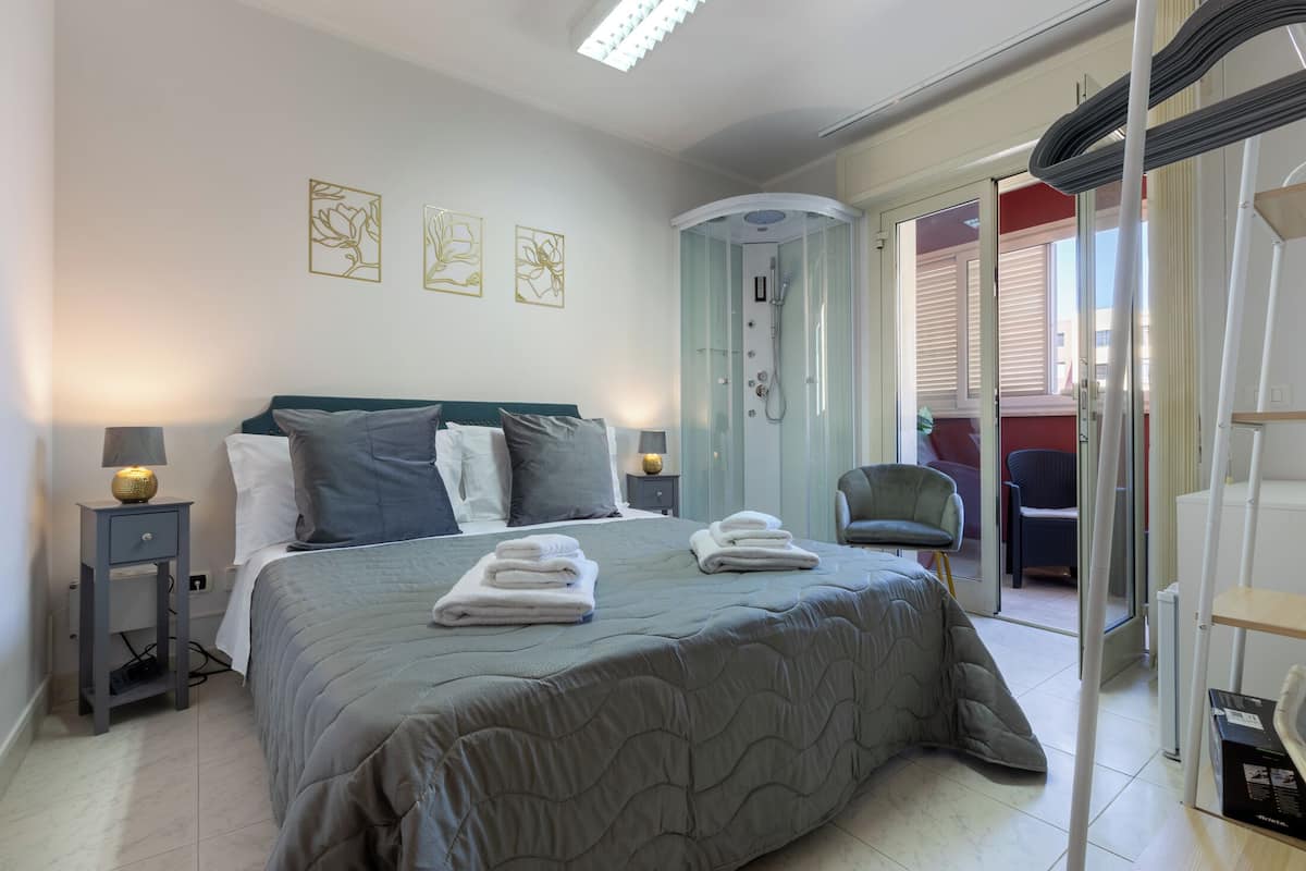 A bedroom with a bed, bedside lamps, a shower, and a chair.