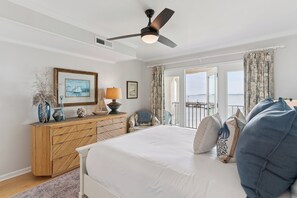 A bedroom with a bed, a ceiling fan, a dresser, a chair, and a view of the sea through a window.