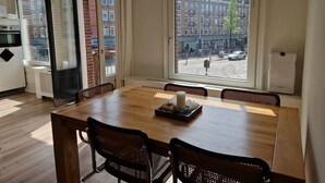 Ruokailutila, jossa on puinen pöytä ja tuolit, ikkuna, josta avautuu näkymä kaupungin kadulle, sekä ovi, joka johtaa keittiöön.