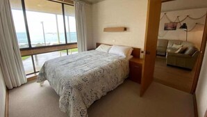 Une chambre à coucher avec un lit, une table de chevet, une étagère en bois, une fenêtre avec des rideaux et une vue sur la mer.