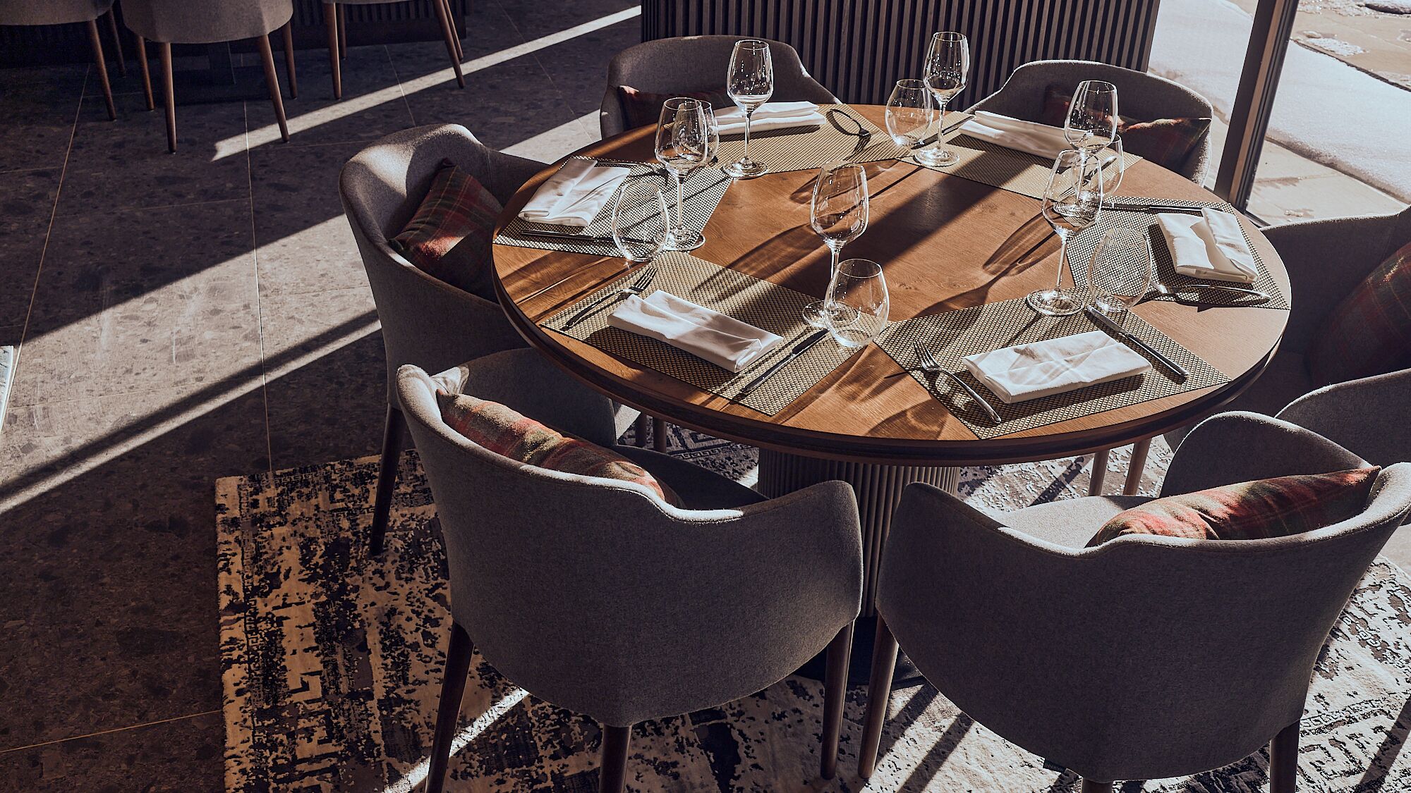 A round dining table with six chairs, each with a plaid cushion. The table is set with wine glasses, cutlery, and folded napkins.