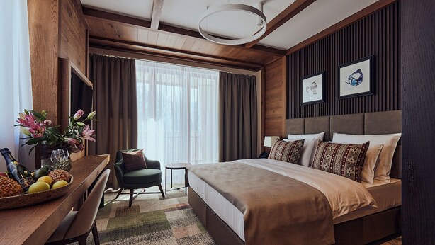 A modern hotel room with a large bed, a desk with a chair, a small table with a fruit basket and a bottle, and a window with curtains.