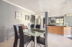 A modern kitchen with a central island, a dining area with a glass table and black chairs, and a grey wall with framed pictures.
