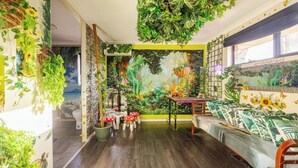 Een kamer met een muurschildering van een tropische tuin, houten vloeren, een bank met een gestreept hoesje, en verschillende potplanten.
