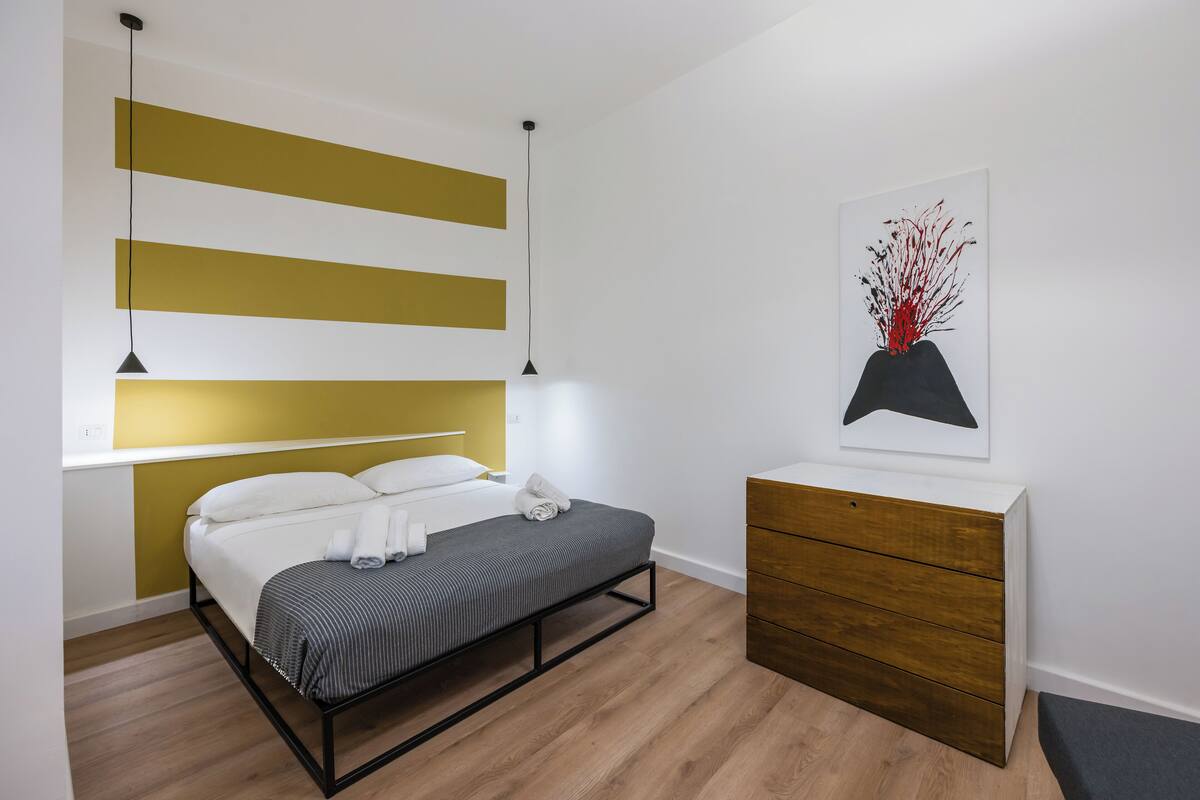 A modern bedroom with a bed, bedside lamps, a wooden dresser, and a wall-mounted shelf.