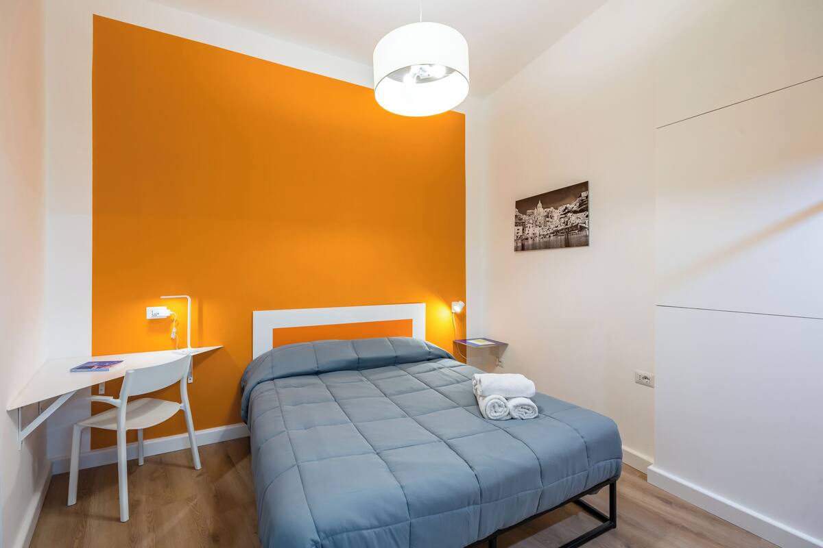 A bedroom with an orange accent wall, a bed with a blue blanket, a white desk with a chair, and a bedside table with a lamp.