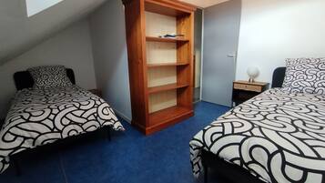 Una habitación pequeña y acogedora con dos camas individuales, una estantería de madera y una mesita de noche con lámpara.
