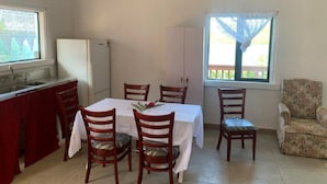 En matplats med ett bord dukat för fyra personer, omgiven av trästolar. I bakgrunden syns ett kylskåp, och det finns ett fönster med utsikt över grönskan.