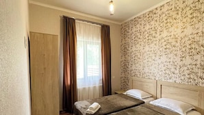 A double bed with two pillows and a folded towel, a wooden wardrobe, a window with curtains, and a patterned wallpaper. - Royal Galaxy

 (Stepantsminda)