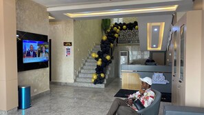 A hotel lobby with a reception desk, a staircase decorated with black and yellow balloons, and a television displaying a news program.