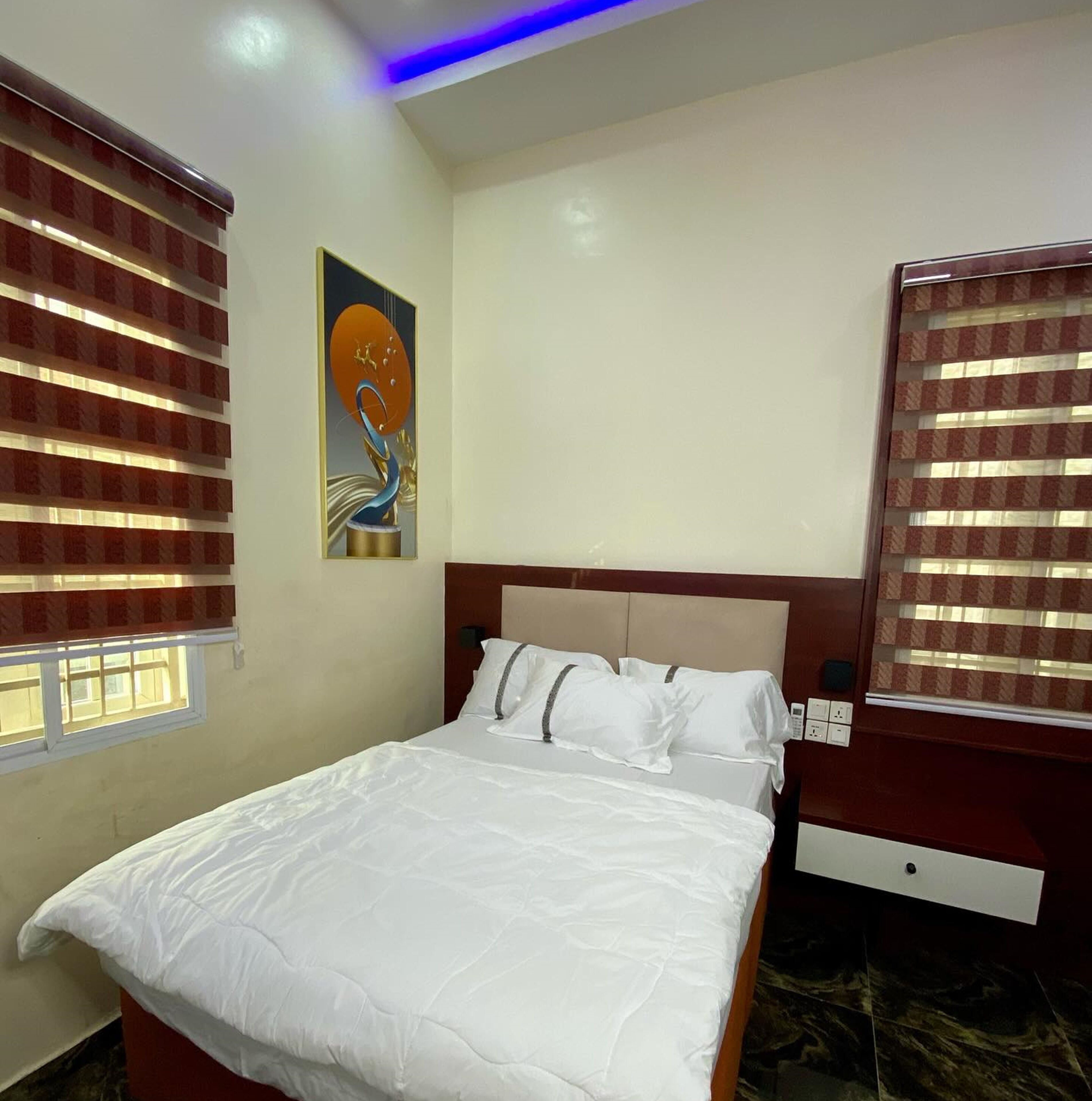 A hotel room with a bed, a nightstand, a wall art piece, and a window with blinds.