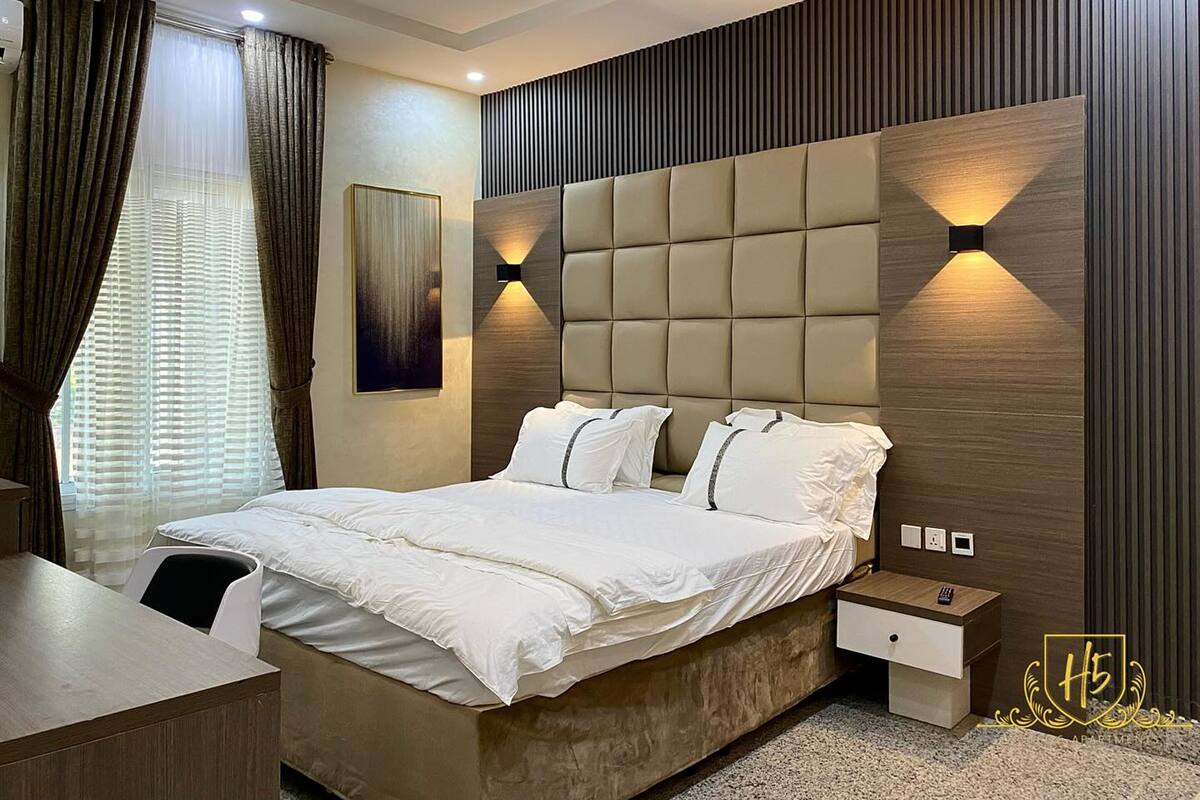 A modern bedroom with a large bed, a bedside table, a desk, and a wall-mounted light fixture.