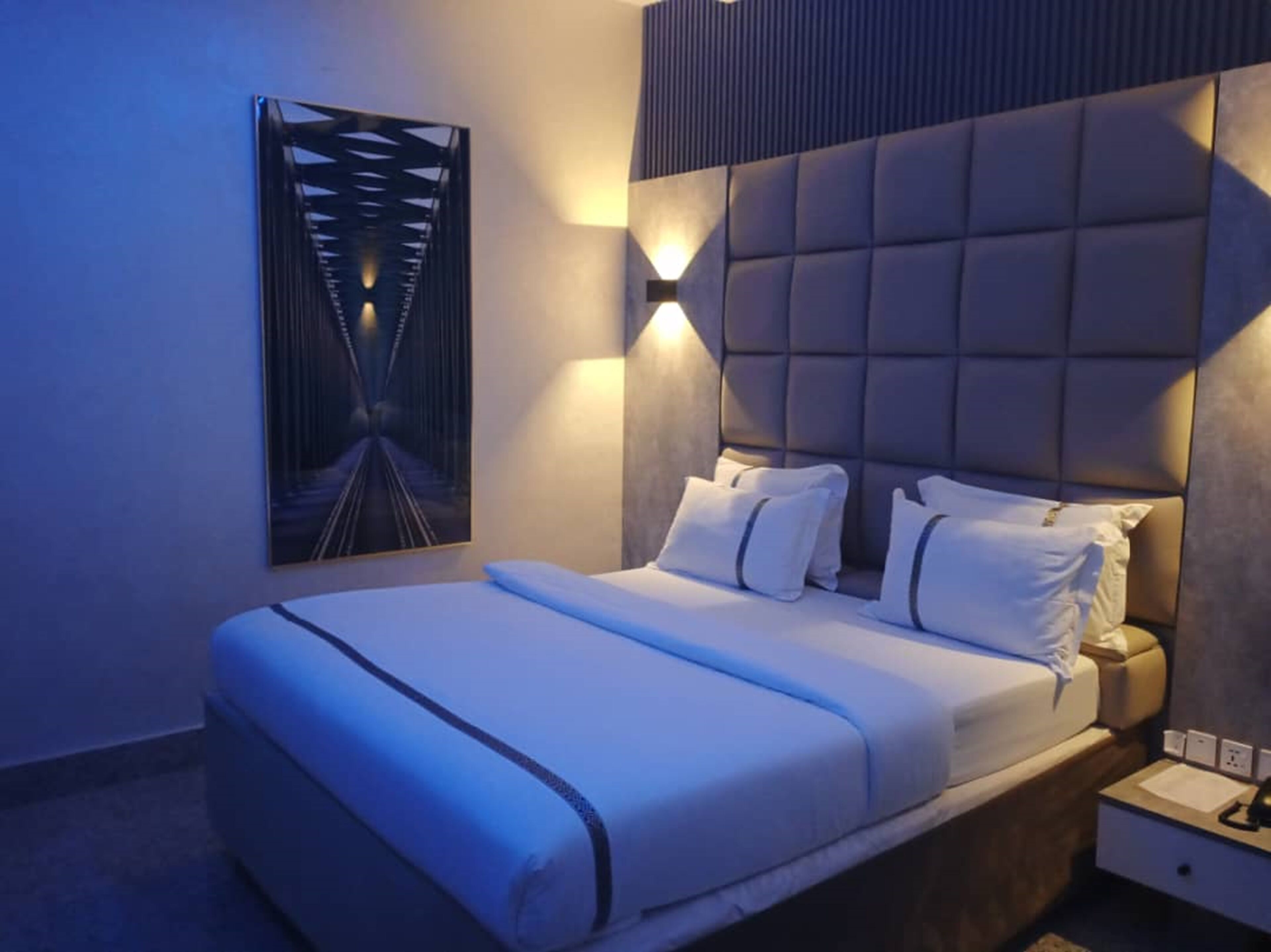 A modern hotel room with a large bed, a headboard with built-in lighting, a nightstand, and a wall-mounted artwork.
