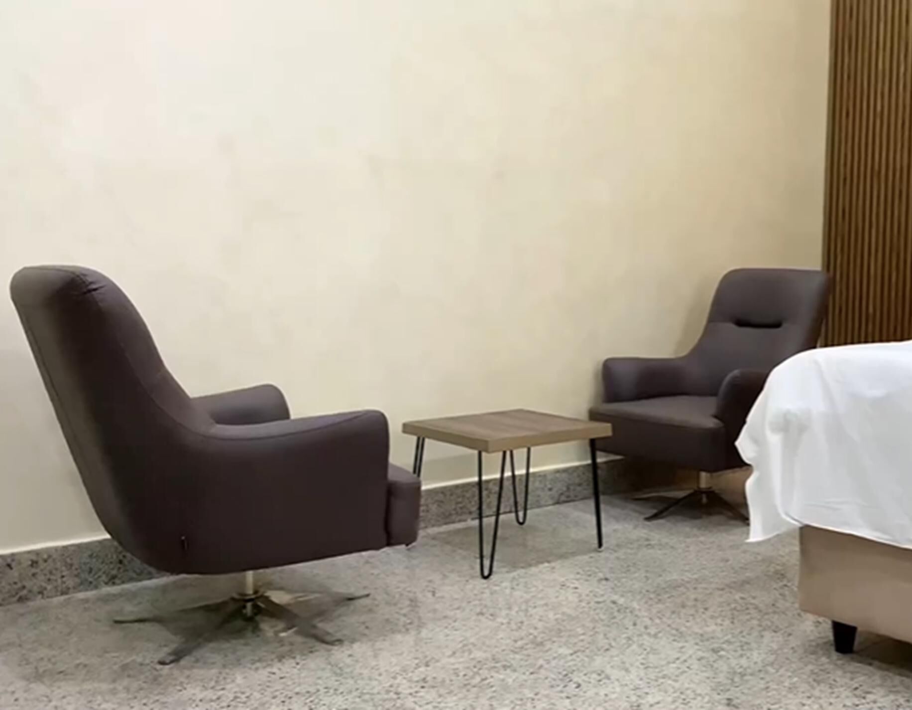 Une pièce comprenant deux fauteuils sombres, une petite table en bois et un lit recouvert d’une couverture blanche.