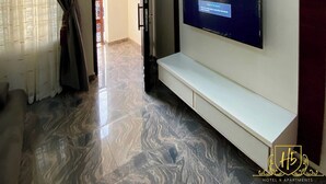 A hotel room with a flat-screen TV mounted on a white wall, a marble-patterned floor, a dark curtain, and a door leading to another room.