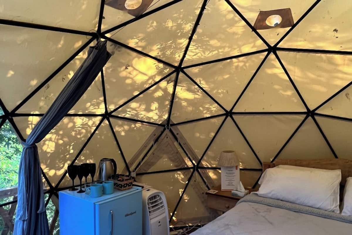 A tent with a geometric ceiling, a bed, a small table with a lamp, and a blue cabinet.