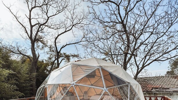 Estrutura em forma de cúpula geodésica com uma cama e uma pequena mesa no interior, rodeada por um deck de madeira e árvores.