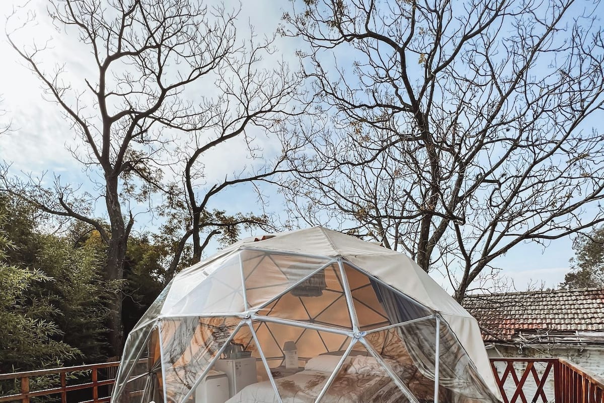A geodesic dome structure with a bed and a small table inside, surrounded by a wooden deck and trees.