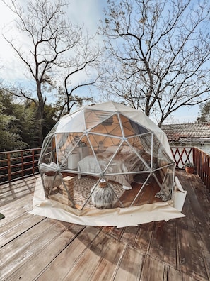 A geodesic dome structure with a bed and a small table inside, surrounded by a wooden deck and trees. - Brisas del Campo Glamping (Zelaya)