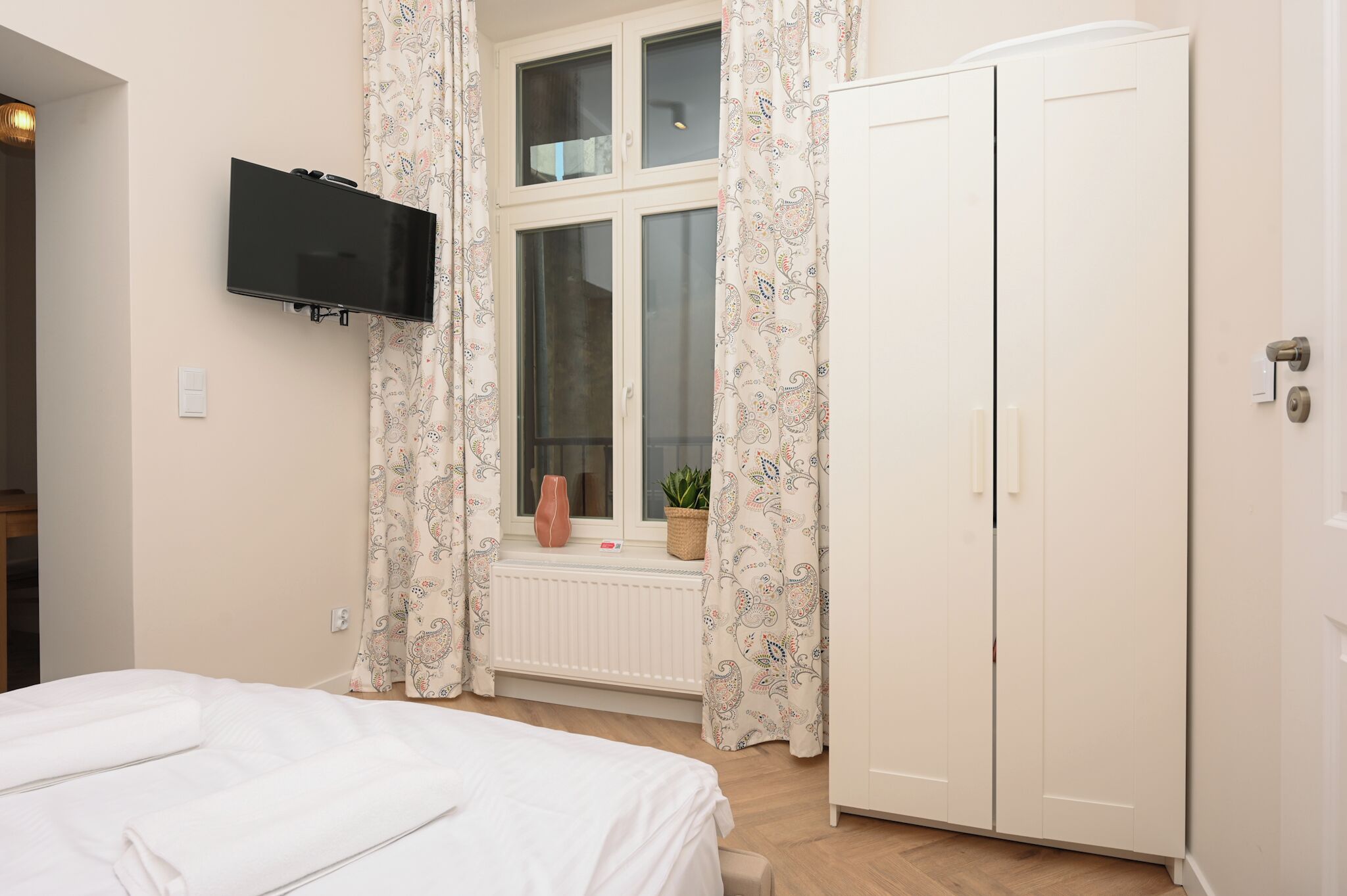 Une chambre à coucher comprenant un lit, un téléviseur fixé au mur, une fenêtre avec des rideaux, un radiateur et une armoire.