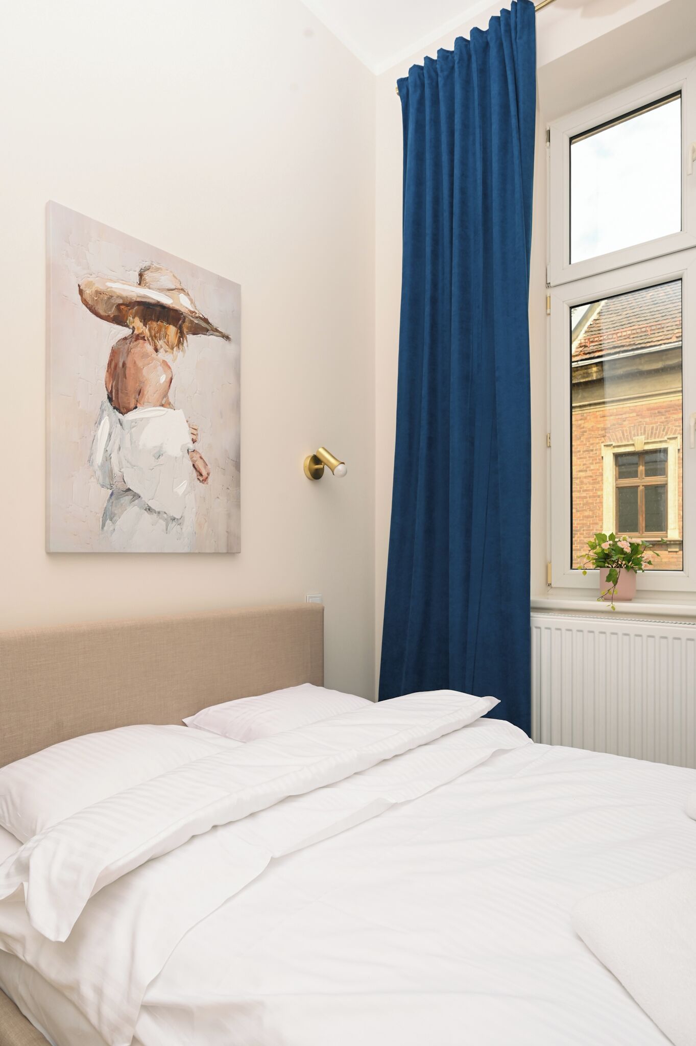 A bedroom with a bed, a painting on the wall, a window with blue curtains, and a view of a building outside.