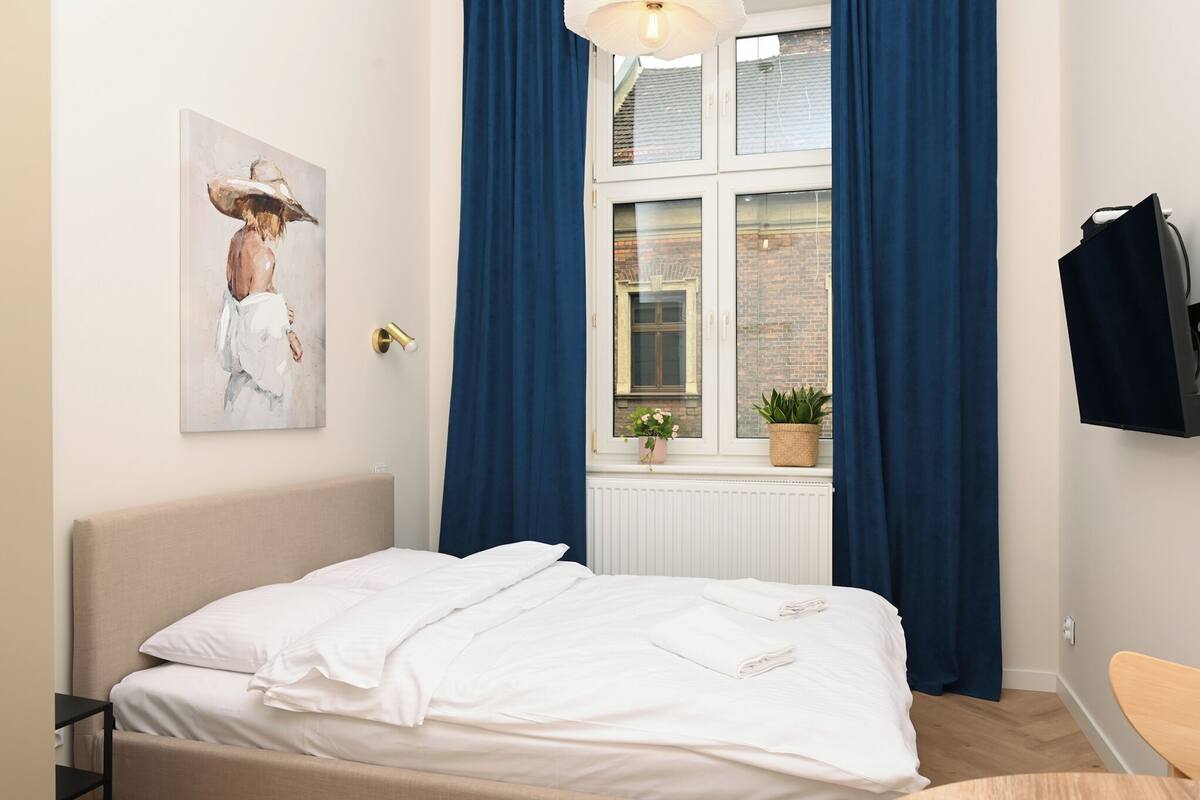 A neatly made bed with white linens, a window with blue curtains, a potted plant on the windowsill, and a painting on the wall.