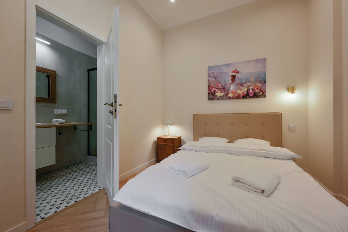 A hotel room with a bed, a nightstand, a painting on the wall, and a bathroom visible through an open door.